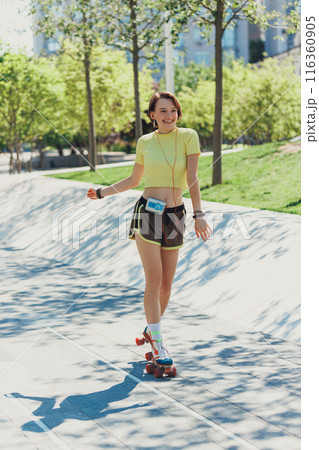 Skater girl training, rolling through park on sunny day, wearing sportswear and retro roller skates, and listening music on vintage player. 116360905
