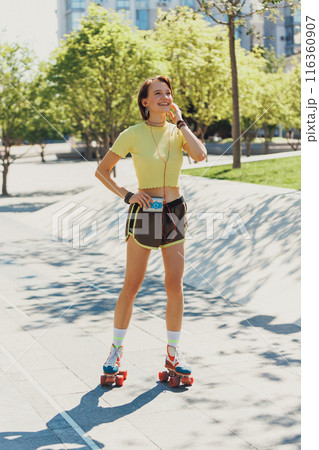Skater girl gleefully rolling through city streets, wearing yellow crop top and brown shorts, and listening music on retro player. 116360907