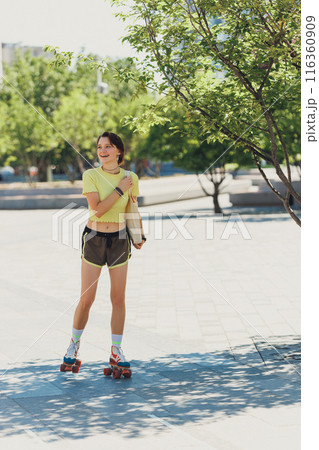 Full-length portrait of energetic young lady in yellow top and sport shorts roller skating with big smile on her face in park. Full-length portrait of energetic young lady in yellow top and sport shorts roller skating with big smile on her face in park. 116360909