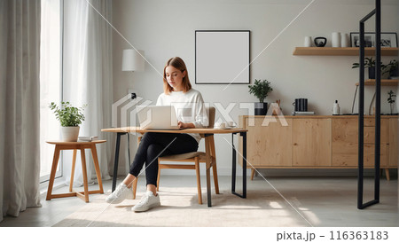 A young woman working on a laptop at a desk in a stylish home office, featuring modern furniture and natural light. 116363183