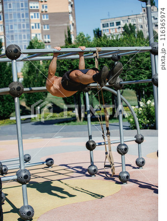 Athletic man in black shorts doing pull-ups on metal frame in park, showcasing muscular body. Early morning training Athletic man in black shorts doing pull-ups on metal frame in park, showcasing muscular body. Early morning training 116365974