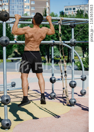Shirtless man performing pull-ups on a metal bar in outdoor fitness park, wearing black shorts. Early morning training Shirtless man performing pull-ups on a metal bar in outdoor fitness park, wearing black shorts. Early morning training 116365983