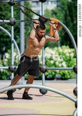 Shirtless man in black shorts working out with suspension straps, doping intense trx exercises in outdoor park, under sunny weather. 116365984
