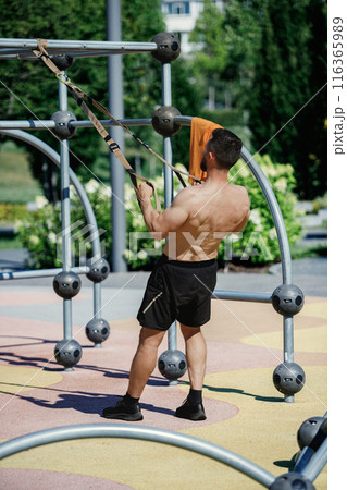 Shirtless man using suspension straps for exercise in outdoor fitness park, wearing black shorts. TRX outdoor workout Shirtless man using suspension straps for exercise in outdoor fitness park, wearing black shorts. TRX outdoor workout 116365989