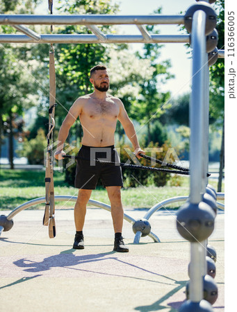 Shirtless man exercising with resistance bands in outdoor park, wearing black shorts, focusing on his workout. Muscular body shape 116366005