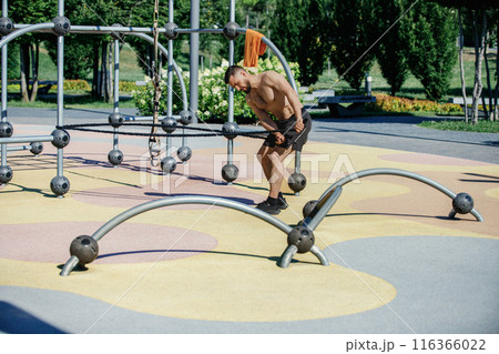Athletic shirtless man training in park, using resistance bands for strength training. Hot morning training session Athletic shirtless man training in park, using resistance bands for strength training. Hot morning training session 116366022