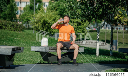 Male fitness enthusiast in park, dressed in bright orange and black, drinking protein drink on bench after intense training. Male fitness enthusiast in park, dressed in bright orange and black, drinking protein drink on bench after intense training. 116366035