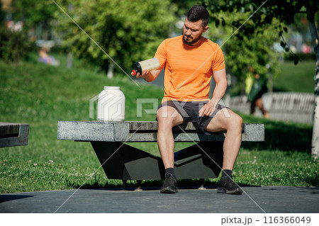 Athletic man wearing orange and black sportswear sitting on bench, using shaker bottle for making proteins shaker. Outdoor training 116366049
