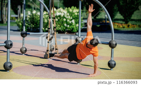 Muscular man performing TRX side plank outdoors, balancing on one arm with his feet suspended. Outdoor intense training 116366093