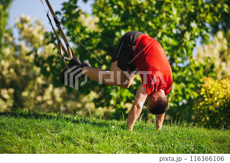 Fit athlete, muscular man in sportswear training outdoors in park, performing an intense TRX pike workout on a grassy ground Fit athlete, muscular man in sportswear training outdoors in park, performing an intense TRX pike workout on a grassy ground 116366106