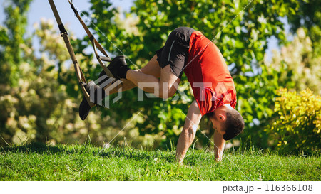 Muscular man doing TRX pike exercise outdoors, with feet suspended in straps. Open air training in park. Muscular man doing TRX pike exercise outdoors, with feet suspended in straps. Open air training in park. 116366108