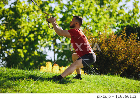Athletic man with song muscular body, in sports clothing training legs with trx fitness straps outdoors against greenery park Athletic man with song muscular body, in sports clothing training legs with trx fitness straps outdoors against greenery park 116366112