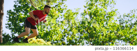 Fit athlete, man in red t-shirt performing TRX exercises outdoors with straps. Muscular man doing squats exercises in greenery park 116366114