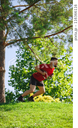 Fit athlete performing TRX exercises outdoors with straps. Muscular man doing squats exercises in greenery park Fit athlete performing TRX exercises outdoors with straps. Muscular man doing squats exercises in greenery park 116366116