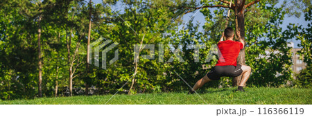Back view. Athletic man in sportswear training outdoors, doing trx workout in greenery park, doing squats exercises. Back view. Athletic man in sportswear training outdoors, doing trx workout in greenery park, doing squats exercises. 116366119