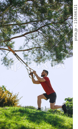 Athletic man doing trx workout outdoors, with hanging straps on tree branches and doing squats. Active and intense training session 116366121
