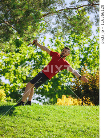 TRX workout session. Muscular bearded man in red t-shirt training outdoors in greenery park, using trx straps for exercises. 116366129