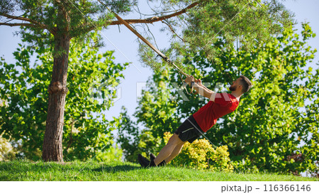 Muscular man doing TRX workout in lush green park, using tree branches for support and demonstrating perfect form and focus. Muscular man doing TRX workout in lush green park, using tree branches for support and demonstrating perfect form and focus. 116366146