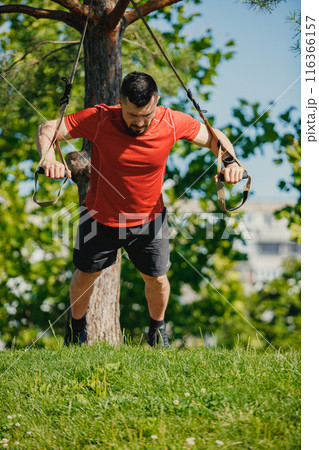 Muscular young man in red shier and black shirt training in the park, doing trx workout. Strong hands and fit body Muscular young man in red shier and black shirt training in the park, doing trx workout. Strong hands and fit body 116366157