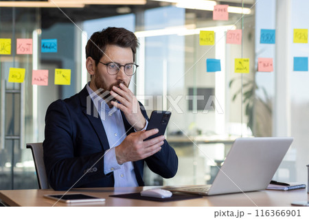 Sad disappointed businessman reading news on phone, man in business suit using app on smartphone, looking upset, received bad news online in email, working with laptop inside office. Sad disappointed businessman reading news on phone, man in business suit using app on smartphone, looking upset, received bad news online in email, working with laptop inside office. 116366901