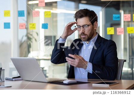 A man in a suit is looking at his cell phone while sitting at a desk. The scene suggests that he might be working or attending to some important matter. The man's facial expression 116366912