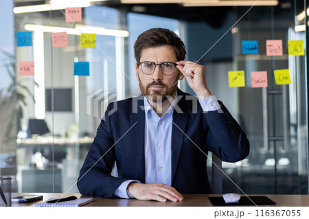 Serious middle-aged businessman deep in thought, sitting in a well-lit modern office, surrounded by sticky notes on glass. Emphasizes strategic planning and decision-making in a corporate environment. 116367055