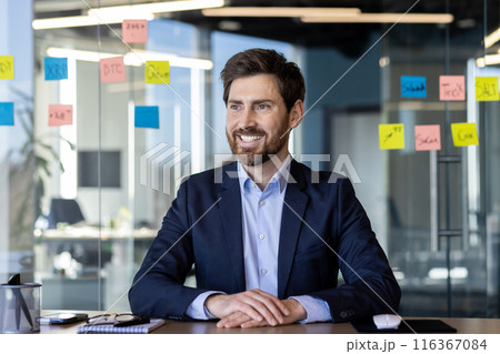 A professional man in a blue suit and white shirt, smiling confidently in a bright, contemporary office setting with visible sticky notes on the glass walls. 116367084