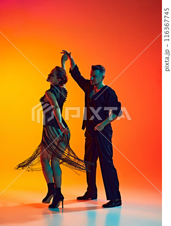 Gentleman and lady, elegant couple dancing bachata, performing with artistry against gradient yellow orange background in neon light Gentleman and lady, elegant couple dancing bachata, performing with artistry against gradient yellow orange background in neon light 116367745