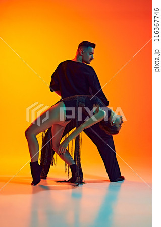 Perfect support. Beautiful young man and woman in black attire dancing passionately bachata against gradient yellow orange background in neon light Perfect support. Beautiful young man and woman in black attire dancing passionately bachata against gradient yellow orange background in neon light 116367746
