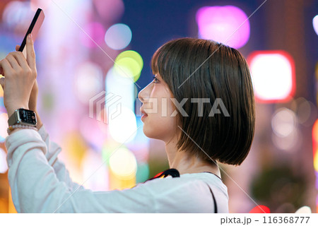 夜の繁華街でスマートフォンをかざす若い女性 116368777