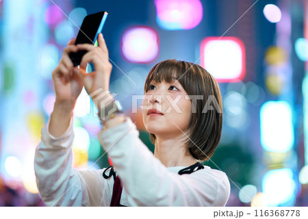 夜の繁華街でスマートフォンをかざす若い女性 116368778