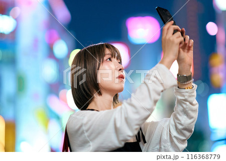 夜の繁華街でスマートフォンをかざす若い女性 116368779