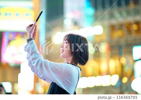 夜の繁華街でスマートフォンをかざす若い女性 116368785