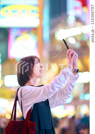 夜の繁華街でスマートフォンをかざす若い女性 116368787