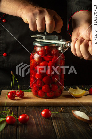 The chef rolls up a jar with a cherry drink or compote with a manual seaming key in his hands. The concept of preserving cherries in jars for future use. Advertising space on the black table 116370713