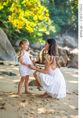 Young woman mother with a little daughter in white dresses on seashore in the shade of trees and palms 116371023