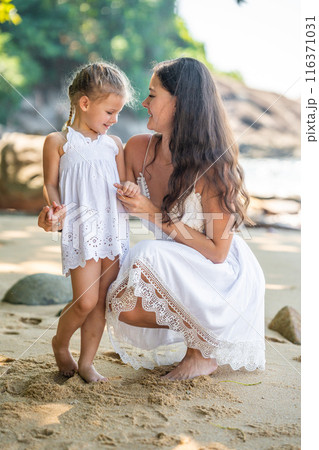 Young woman mother with a little daughter in white dresses on seashore in the shade of trees and palms Young woman mother with a little daughter in white dresses on seashore in the shade of trees and palms 116371031