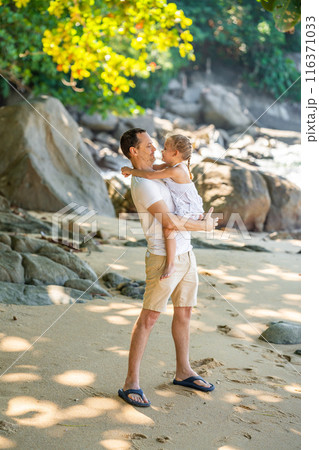 Little girl on father's hands smile and rejoice on seashore in the shade of trees and palms. Happy family concept 116371033