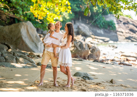 Happy young family of three taking a walk on the beach, having a fun on seashore in the shade of trees and palms. Happy family concept. Happy young family of three taking a walk on the beach, having a fun on seashore in the shade of trees and palms. Happy family concept. 116371036