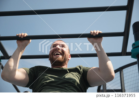Handsome man does pull-ups on horizontal bar at street on sports ground. Outdoors. Summer. Workout. Morning routine. Handsome man does pull-ups on horizontal bar at street on sports ground. Outdoors. Summer. Workout. Morning routine. 116371955