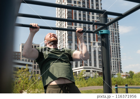Handsome bearded man does pull-ups on horizontal bar at street on sports ground. Outdoors. Summer. Workout. Morning routine. Handsome bearded man does pull-ups on horizontal bar at street on sports ground. Outdoors. Summer. Workout. Morning routine. 116371956
