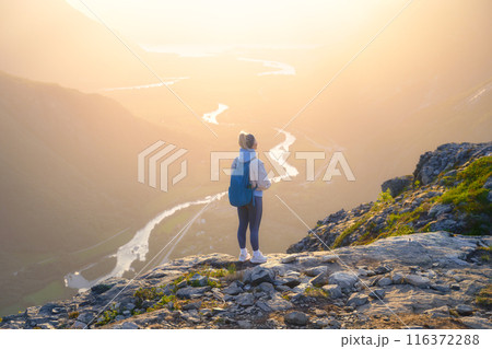 A young athletic girl standing on a rock with a backpack. Sunshine in a mountain valley. 116372288