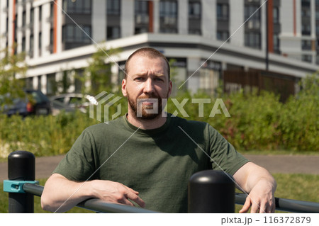 Portrait of caucasian handsome bearded man wearing military color t-shirt. Athlete man on outdoor sports ground. Courtyard. Outdoors. Summer. 116372879