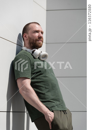 Portrait of caucasian handsome beard man wearing military color t-shirt with headphones near modern white wall listening music. Favourite song. Copy space. 116372880