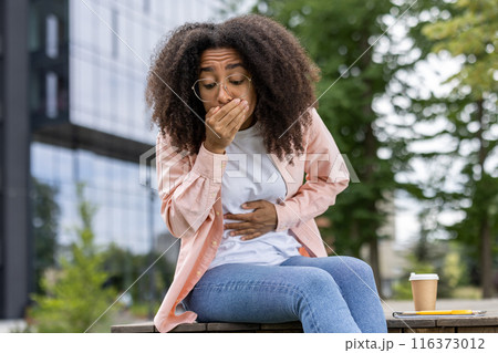 Young woman experiencing stomach pain and holding her hand over mouth. She is sitting outside in casual attire, indicating discomfort and possible nausea. Young woman experiencing stomach pain and holding her hand over mouth. She is sitting outside in casual attire, indicating discomfort and possible nausea. 116373012