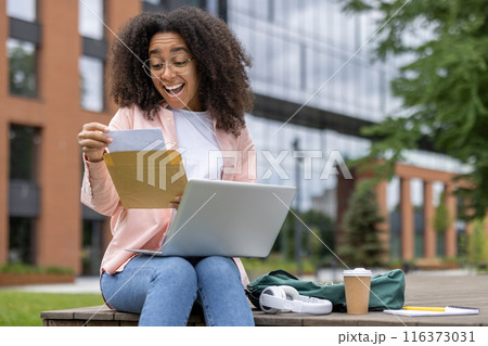 Happy student receives letter of approval sitting on university campus bench with laptop. Joyful reaction to good exam results or enrollment confirmation while studying outdoors. Happy student receives letter of approval sitting on university campus bench with laptop. Joyful reaction to good exam results or enrollment confirmation while studying outdoors. 116373031