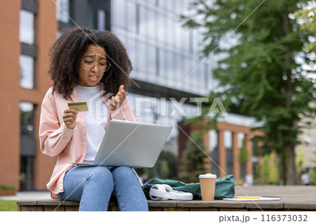 Woman with curly hair sitting outdoors holding credit card, looking worried while using laptop. Concept of online shopping, financial issues, and remote work. Woman with curly hair sitting outdoors holding credit card, looking worried while using laptop. Concept of online shopping, financial issues, and remote work. 116373032