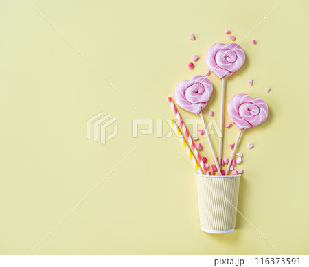 Pink lollipops in the shape of hearts at  paper cup with color sugar on a yellow background.  116373591