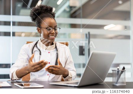 Professional doctor sitting at desk conducting video call consultation in modern office. Stethoscope around neck, gestures to communicate effectively. Medical professional using technology Professional doctor sitting at desk conducting video call consultation in modern office. Stethoscope around neck, gestures to communicate effectively. Medical professional using technology 116374040