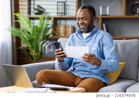 Smiling man working remotely from home, using smartphone and reviewing document while sitting on sofa with laptop and notepad. Concept of remote work home office, happiness, productivity, technology. 116374056